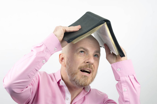 Γενειοφόρος άνδρας σε ένα ροζ πουκάμισο κρύβει το κεφάλι του κάτω από μια ανοικτή Αγία Γραφή - Φωτογραφία, εικόνα