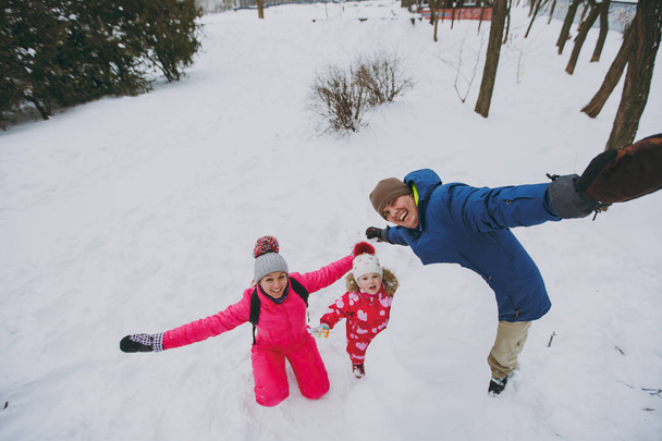Забавный семейный мужчина, женщина и маленькая девочка в теплой одежде играют, заставляя снеговика расправлять руки в парке или лесу на открытом воздухе. Зимнее веселье, отдых в праздники. Семейный стиль жизни
 - Фото, изображение