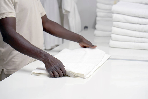 Hands of an african male laundry hotel worker folds a clean white towel. Hotel staff workers. Hotel linen cleaning services. - Zdjęcie, obraz