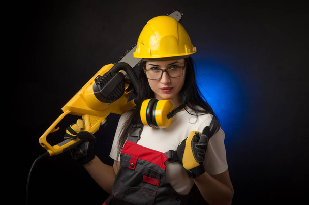 brunette girl in special clothes and a worker in a helmet posing on a black background with a working tool - Foto, Imagen