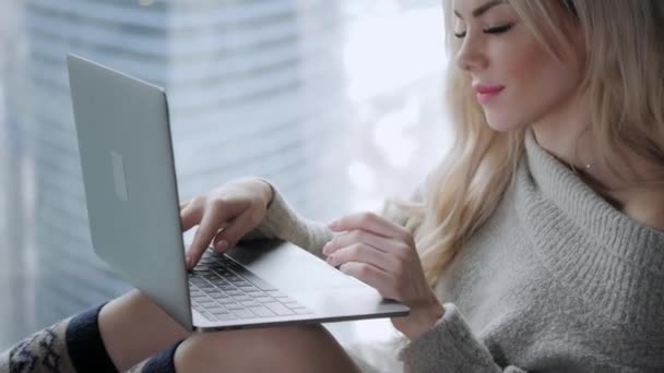 Beautiful blonde woman sitting on the bed with gray aluminum laptop. Girl smiles, good mood. Blogging, browsing internet, chatting. In a warm cozy sweater and wool socks. Happy winter concept. - 映像、動画