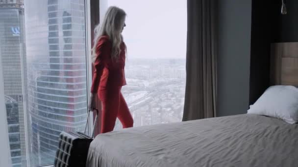 Joven mujer de negocios rubia llega a una habitación de hotel con maleta negra. Mujer con traje de negocios de coral rojo. Chica joven entra en la habitación sosteniendo la maleta por el mango
 - Imágenes, Vídeo