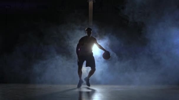 ein Mann mit einem Basketball auf einem dunklen Basketballfeld vor dem Hintergrund eines Basketballrings im Rauch zeigt Dribbelkünste, die von drei Laternen im Gegenlicht beleuchtet werden - Filmmaterial, Video