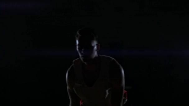 Male urban basketball player dribbles ball in crouched position in an inner-city basketball court lit by single street light - Filmati, video