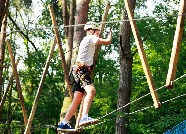 Симпатичный мальчик, наслаждающийся активностью в парке скалолазания в солнечный летний день. Ребенок залезает на верёвочную площадку. Безопасное восхождение со страхованием шлема. Детский парк приключений в лесу, экстремальный спорт
 - Фото, изображение