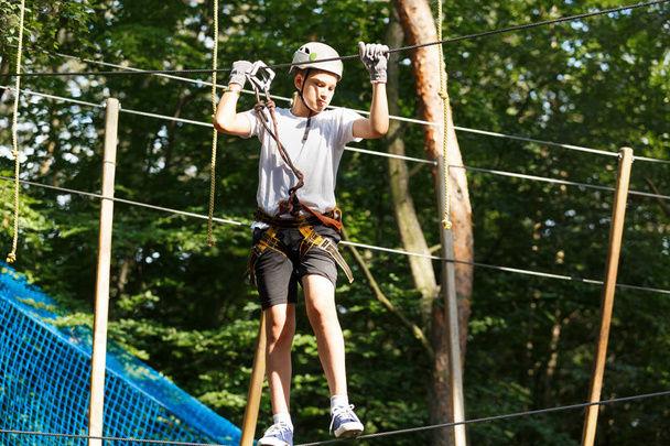 Cute boy enjoying activity in climbing adventure park at sunny summer day. Kid climbing in rope playground structure. Safe climbing with helmet insurance. Child in forest adventure park, extreme sport - Photo, Image