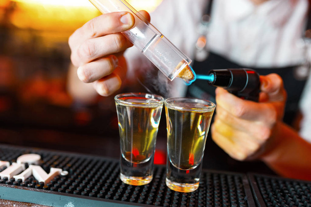 Bartender man making hot alcohol shots on the bar in the pub with a professional burner. The bartender lights a lighter over a glass.Relax in the nightclub. Hot fire drinks. Lets the party - 写真・画像