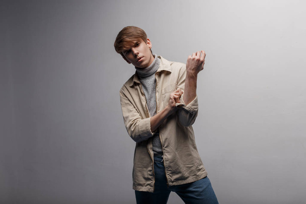 stylischer amerikanischer junger Mann in leichter modischer Jacke in grauem Strickgolf in blauer modischer Hose mit modischer Frisur, der im Studio vor einer weißen Wand posiert. attraktiver Typ - Foto, Bild