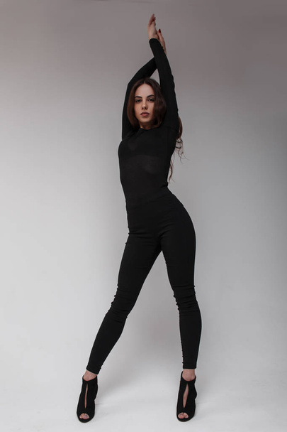 Jeune femme sexy attrayante avec une belle silhouette dans un T-shirt noir à la mode en leggings élégants noirs et des chaussures noires avec des talons posant dans un studio lumineux contre un mur blanc. Magnifique fille
 - Photo, image
