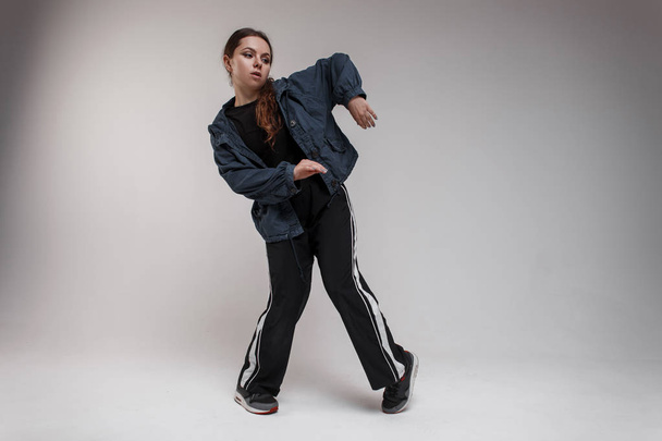 Attractive young woman dancer with brown long hair in fashionable black sportswear demonstrates the movements of the dance to the camera indoors near a white wall. Modern choreography - Foto, immagini