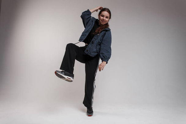 A talented woman dancer in a blue jacket in a stylish T-shirt in fashionable black sports pants is standing on one leg in the studio for white background. Stylish hipster girl dancing - Фото, зображення
