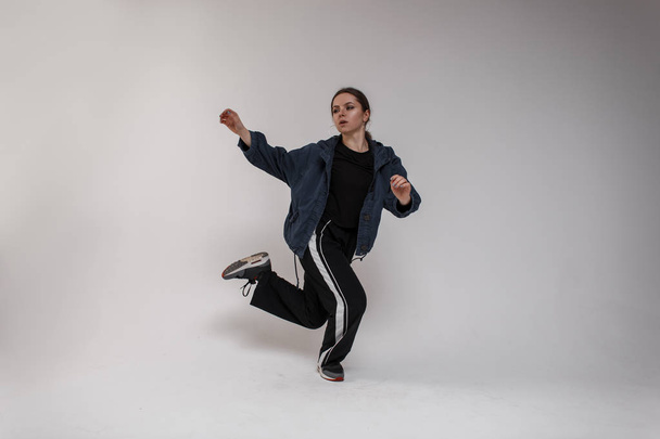 Pretty young woman dancer in a denim jacket in a black stylish T-shirt in sports pants dancing in a bright studio against a white wall. Sports girl - Foto, immagini