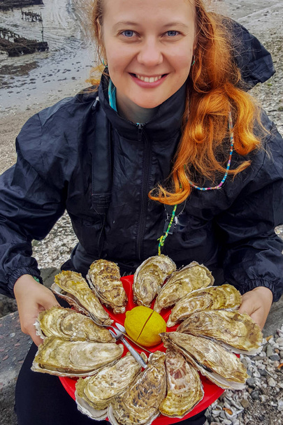 Kaukaski młody biała kobieta z rude włosy trzyma w dłoniach płytkę z ostryg i cytryny na tle Morze Celtyckie - Zdjęcie, obraz