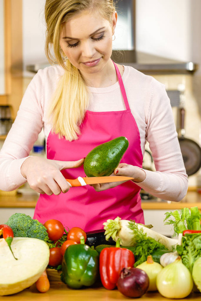 Woman cutting, preparing green vegetable, delicious avocado using kitchen knife. Female wearing apron making food. - Foto, Bild