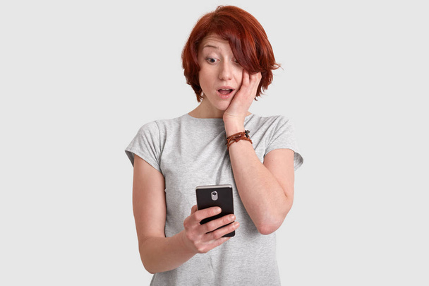Photo of stupefied red haired woman in casual outfit holds mobile phone, recieves unexpected notification, concentrated in screen, isolated over white background. People and reaction concept - Фото, изображение