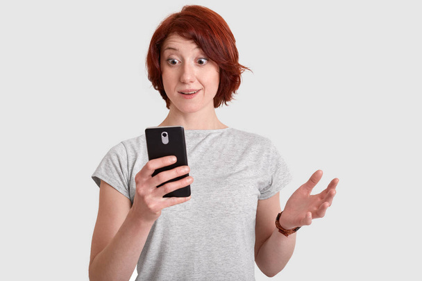 Indoor shot of surprised redhead woman with short hair, spreads hand, reads information from website, has wondered expression, holds mobile phone, isolated over white background, types text message - Valokuva, kuva