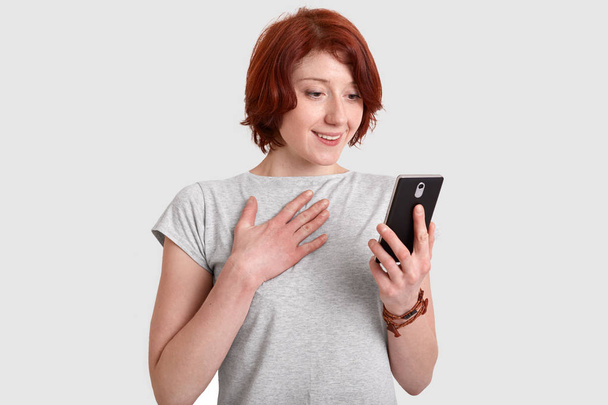 Blij lachende rood harige vrouw houdt hand op de borst, wachtruimten mobiele telefoon, glimlacht zachtjes, ontvangt bericht van felicitatie van beste vriend, gekleed in een casual t-shirt, modellen op witte achtergrond - Foto, afbeelding