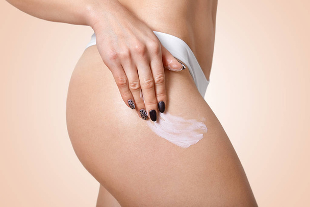 Sideways shot of slim woman applies body cream on hip, wants to have flawless ideal skin, has black manicure, wears white underwear, isolated over beige background. Lady uses lotion for legs. - Zdjęcie, obraz
