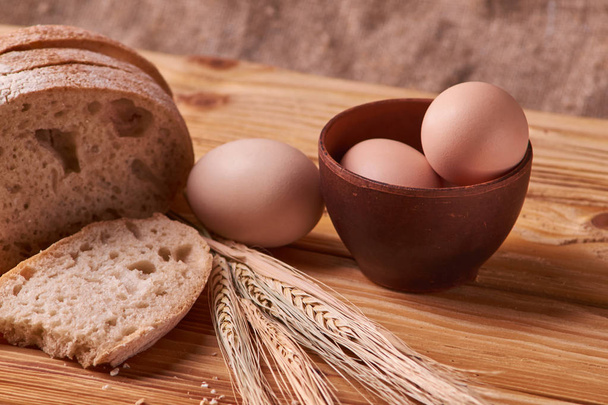pão delicioso fresco, conceito para assar. Ovos em tigela de barro e brotos de trigo na mesa de madeira
 - Foto, Imagem