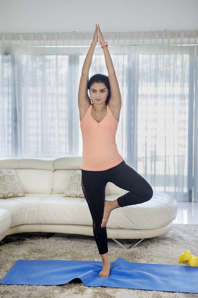 Full length of curly hair woman wearing sportswear while exercising yoga with tree pose at home - Foto, imagen