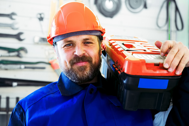 Un trabajador en un casco con una caja de herramientas en el hombro mira directamente a la cámara y sonríe. Hombre barbudo aspecto caucásico en la ropa de trabajo en el fondo de la pared con herramientas
. - Foto, imagen