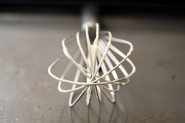 metal wire whisk with dough leftovers on a grey baking tray, close up view with copy space, selected focus and very narrow depth of field - Photo, Image