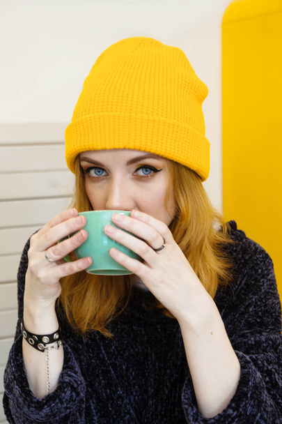 Młoda kobieta o niebieskich oczach i blond włosy w kapelusz żółty dziewiarskich jest picie kawy w kawiarni. - Zdjęcie, obraz