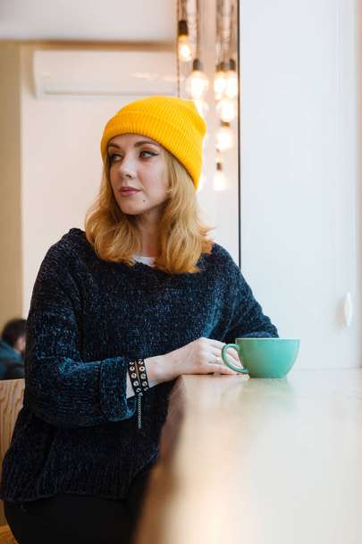Retrato de mujer joven con ojos azules y cabello rubio en un sombrero de punto amarillo está bebiendo café en un café cerca de una ventana
. - Foto, imagen