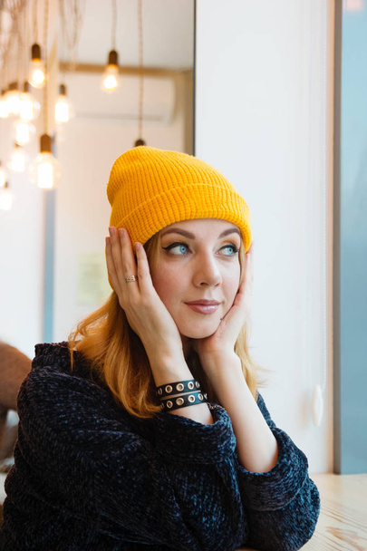 Young woman with blue eyes and blond hair in a yellow knitting hat is sitting in a cafe and looking into the window. - Foto, imagen