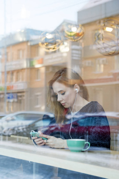 Młoda kobieta z blond włosy i niebieskie oczy z słuchawki, smartphone i filiżankę kawy jest siedząc w kawiarni za oknem. - Zdjęcie, obraz
