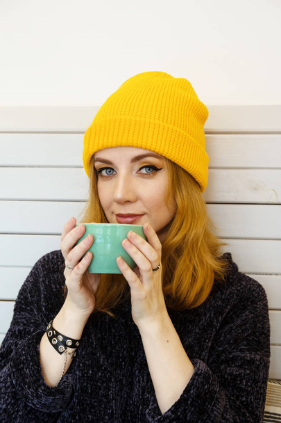 Young woman with blue eyes and blond hair in a yellow knitting hat is drinking coffee in a cafe. - Fotografie, Obrázek