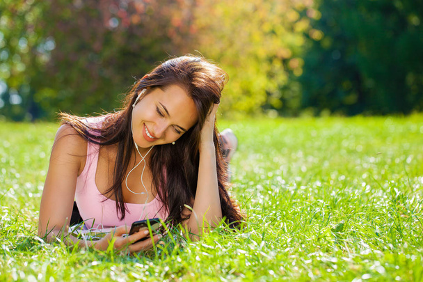 Young beautiful woman in dress lies on green grass and listens to music in headphones through smartphone. - Fotó, kép