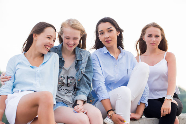 Four attractive women in summer clothes sitting at concrete border near river after studing in college. Young student girls having fun - Photo, Image