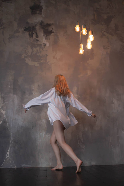 schöne schlanke junge Balletttänzerin im modernen Jazz-Stil, die ein schwarzes Trikot und ein weißes Hemd auf einem Loft-Studio-Hintergrund trägt - Foto, Bild