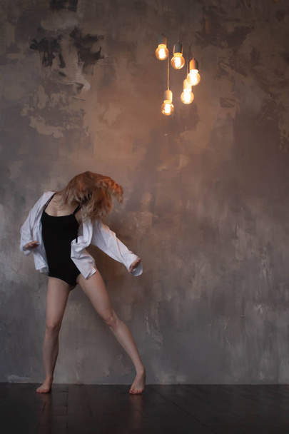 Hermosa joven y delgada bailarina de ballet de estilo contemporáneo de jazz femenino con un maillot negro y una camisa blanca sobre un fondo de estudio tipo loft
 - Foto, Imagen