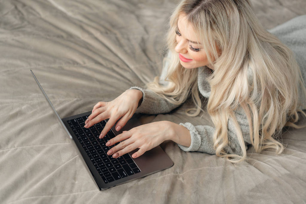 Belle femme blonde allongée sur le lit et regarde l'écran de l'ordinateur portable. Bloguer, naviguer sur Internet, bavarder. Dans un pull chaleureux et des chaussettes en laine
. - Photo, image