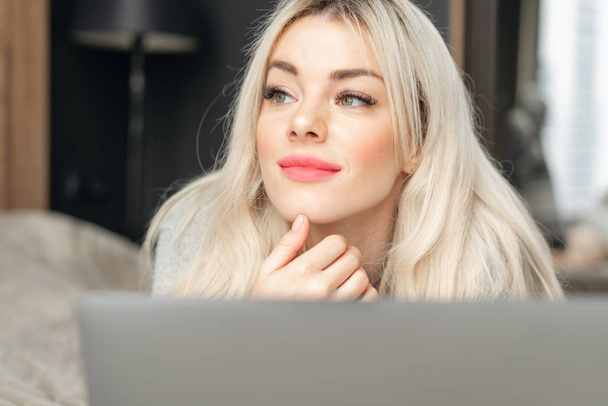 portrait of a girl behind a laptop screen. Beautiful blonde woman lying on the bed and looks at the laptop screen. Blogging, browsing internet, chatting. In a warm cozy sweater - Photo, Image