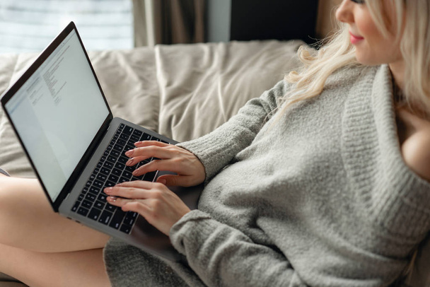 Beautiful blonde woman lying on the bed and looks at the laptop screen. Blogging, browsing internet, chatting. In a warm cozy sweater and wool socks. - Photo, Image