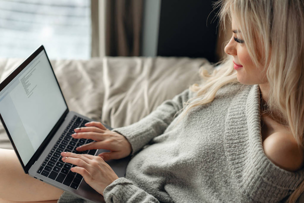 Красива блондинка лежить на ліжку і дивиться на екран ноутбука. Блог, перегляд інтернету, балачки. У теплому затишному светрі і вовняних шкарпетках
. - Фото, зображення