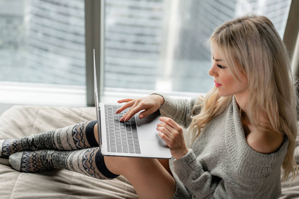 Κορίτσι δακτυλογράφηση στο πληκτρολόγιο. Όμορφη ξανθιά γυναίκα που κάθεται στο κρεβάτι και κοιτάζει την οθόνη του φορητού υπολογιστή. Blogging, περιήγηση στο internet, κουβεντιάζοντας. Σε έναν ζεστό άνετο πουλόβερ και κάλτσες μαλλιού. - Φωτογραφία, εικόνα