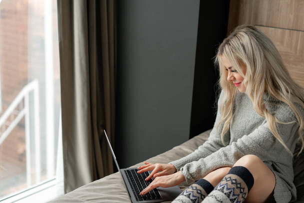 schöne blonde Frau sitzt auf dem Bett mit grauem Aluminium-Laptop. Mädchen lächelt, gute Laune. Bloggen, surfen im Internet, chatten. in einem warmen kuscheligen Pullover und Wollsocken. Frohes Winterkonzept. - Foto, Bild