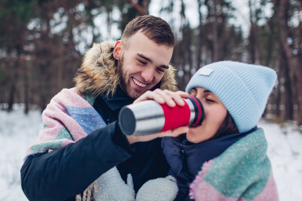 Homme donnant à sa petite amie du thé chaud à boire dans une tasse thermos. Couple amoureux marchant dans la forêt d'hiver couvert de couverture
 - Photo, image