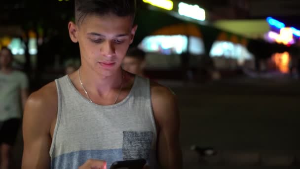 Thoughtful young man browsing the net on his phone in a city at night in summer                      Portrait of a bright brunet guy with short haircut in a grey a-shirt looking at photos on his smartphone in a city street at night in summer - Footage, Video