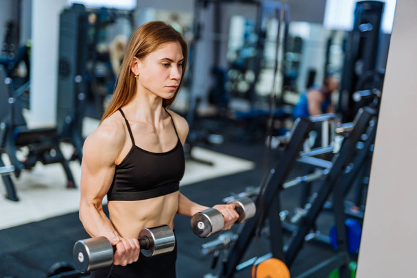 Strong athletic woman in black sport wear pumping up muscles with dumbbells in the gym. Fitness girl with perfect body in the gym has a training. - Foto, Bild