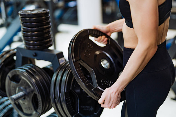 Athletic young woman holding disc from barbell and exercising in gym. Woman's hands taking barbell disk on the sports equipment background. Close-up - Foto, afbeelding