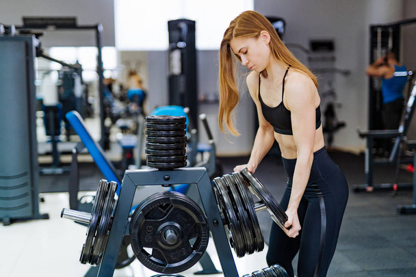 Beautiful strong young sports woman puts barbell disk at fitness gym. Active fitness female with barbell plates in gym. - Valokuva, kuva