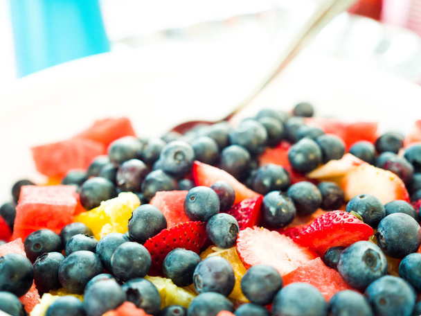 Fotografía de cerca de una ensalada de frutas hecha en casa para una fiesta de género revelan que contiene arándanos, fresas, piña y sandía en un tazón de servir blanco y decoración azul y rosa más allá
. - Foto, imagen