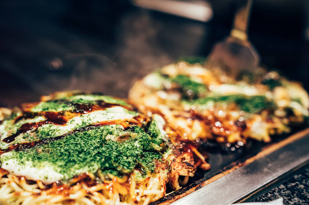 cooking okonomiyaki in local restaurant on teppanyaki pan. delicious Japanese hot plate pizza with green vegetable and sauce on it surrounding by smoke on iron plate indoor. - Fotoğraf, Görsel