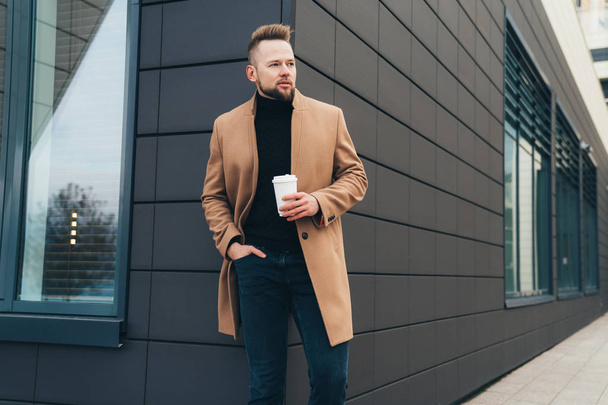 Businessman in brown coat and cup of coffee in his hands posing on background of modern building - Foto, Imagem