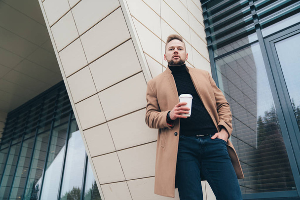 Attractive man in trendy brown coat and coffe in his hand posing near business center - Foto, Imagen
